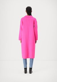 Lång rosa ullkappa med krage och långa ärmar, med rak skärning, i kombination med ljusblå jeans och svarta klumpiga stövlar.