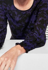 Maglietta a maniche lunghe in viola e nero con un motivo floreale, caratterizzata da maniche arricciate e un colletto rotondo, realizzata in tessuto morbido.