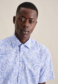 Camicia a maniche corte con bottone in tessuto azzurro chiaro con un motivo paisley bianco. Presenta un colletto con punte e un dettaglio di tasca anteriore.