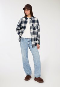 Camicia flanella a quadri in blu navy e bianco, abbinata a una maglietta bianca semplice e jeans azzurri chiari. Scarpe slip-on in suede marrone e un cappellino scuro.