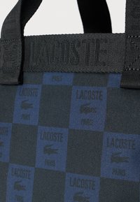 Czarny i niebieski materiał w kratkę z powtarzającym się logo Lacoste i emblematem krokodyla, wyposażony w czarną teksturowaną rączkę z napisem „Lacoste”.
