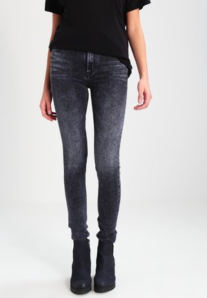 Strak zittende zwarte jeans met acid-wash, gedragen met zwarte enkellaarzen en een zwart kortärmelig topje, staand tegen een effen lichte achtergrond.