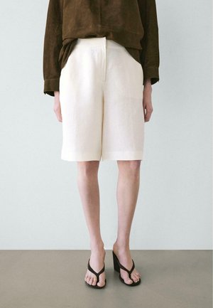 Personne portant un short blanc jusqu'aux genoux, un haut à manches longues marron foncé et des sandales tongs noires à talons hauts, debout sur un sol gris.