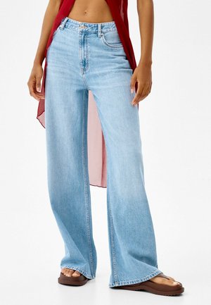 Jeans de mezclilla de pierna ancha de color azul claro con una cintura alta, que presentan un cierre de botón, grandes bolsillos y un ajuste holgado. Combinados con sandalias de dedo marrones.
