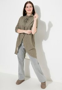 Cardigan lungo verde oliva con collo a cravatta, spacchi laterali e chiusura a bottoni, abbinato a jeans grigi chiari a zampa d'elefante e scarpe in camoscio marroni.
