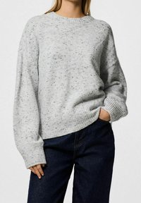 Femme portant un pull ample en maille gris clair moucheté avec des poignets côtelés et un jean taille haute bleu foncé, main dans la poche.