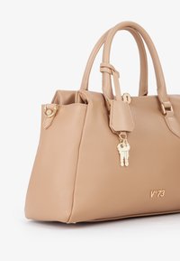 Beige leren handtas met dubbele handgrepen, goudkleurige hardware, sleutelhanger detail en gecursiveerd logo. Getextureerd oppervlak en gestructureerde vorm.