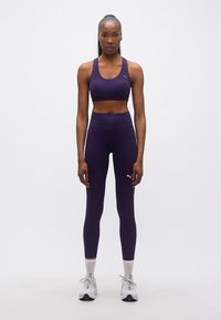 Γυναίκα που στέκεται όρθια φορώντας αθλητικό σουτιέν σκούρου μοβ και κολάν με λευκά αθλητικά παπούτσια σε λευκό φόντο.