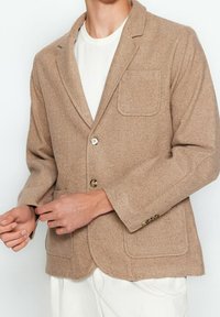 Blazer beige in lana con rever a punta, chiusura con un solo bottone, due tasche frontali e una finitura testurizzata. Camicia bianca sotto.