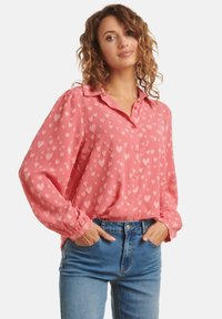 Blouse rose avec motifs en cœur, col boutonné et manches froncées, portée avec un jean bleu. Tissu léger, coupe décontractée et texture lisse.
