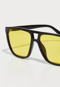 Ochelari de soare negri din plastic, cu rame pătrate și lentile galbene. Detalii notabile sub formă de decupaj între lentile și brațe. Textură netedă, design modern.