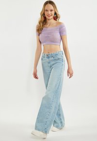 Fialový pletený top s odhalenými rameny zkráceného střihu doplněný o světle modré široké džíny. Model má na sobě bílé tenisky a barevné visací náušnice.