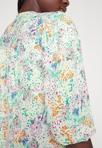 Blusa de padrão floral em verde claro com mangas bufantes curtas. Apresenta detalhes florais em rosa, laranja e azul em tecido leve.