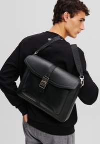 Borsa a tracolla in pelle nera con patta pieghevole, tracolla regolabile con motivo e dettagli in metallo. Texture liscia con forma strutturata.