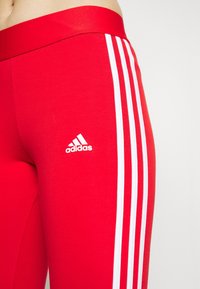 Czerwone legginsy sportowe z elastycznego materiału, z trzema białymi paskami po bokach i białym logo Adidas na udzie.