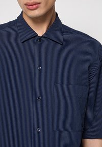 Camisa de manga corta en tela azul marino con rayas, con cuello puntiagudo, cierre frontal con botones, un bolsillo en el pecho y rayas texturizadas.