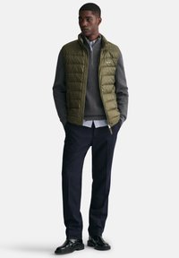 Chaleco de plumas color caqui sobre un suéter gris, combinado con pantalones marinos y zapatos negros. Presenta un cuello alto y un cierre frontal con cremallera.