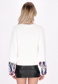 Maglione bianco e soffice con maniche ampie che presenta accenti di paillettes colorate in rosso, blu e argento sui polsini. Sotto una gonna di pelle nera.