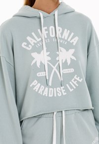 Lichtblauwe cropped hoodie van zacht materiaal, met een witte grafische print van palm bomen en de tekst "CALIFORNIA PARADISE LIFE."