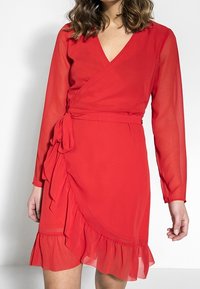 Vestido envolvente rojo con mangas largas de tela transparente, escote en V, cinturón en la cintura, bajo con volantes y textura de tela fluida.