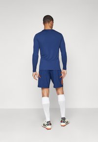 Maglietta sportiva blu a maniche lunghe abbinata a pantaloni corti blu con dettagli arancioni. Calze bianche al ginocchio e scarpe sportive grigie completano l'outfit.