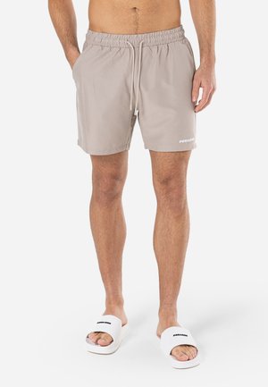 Shorts de bain beiges avec une taille élastique, un cordon de serrage et des poches latérales. Détails du logo blanc sur la jambe. Portés avec des sandales blanches.