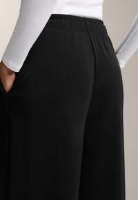 Zwarte wijde broeken hebben een elastieken tailleband en zijzakken, gemaakt van een gladde, rekbare stof met een matte afwerking.