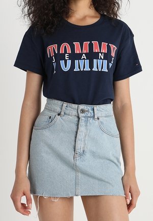 Kvinde iført en navy blå Tommy Jeans t-shirt, som er stoppet ned i et lyseblåt denim miniskørt med frynset kant.