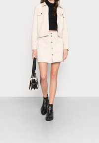 Beige kordjacka med två bröstfickor, i kombination med en matchande knappförsedd kjol. Svarta anklarstövlar med cirkulär hårdvara, bär en svart väska.