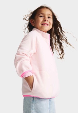 Jasnoróżowy polarowy pulower z wysokim kołnierzem, falistymi różowymi mankietami i krawędzią. W zestawie boczne kieszenie i zamek błyskawiczny dla łatwego zakładania.