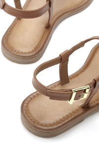 Braune Leder-Sandalen mit verstellbaren Riemen, flacher Sohle und goldener Schnallendetail. Glatte Oberfläche mit einer Naht, die die Kanten umreißt.