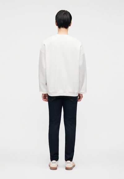 Λευκή φαρδιά φούτερ μπλούζα με χαλαρούς ώμους, συνδυασμένη με σκούρα μπλε στενά τζιν και μπεζ αθλητικά παπούτσια. Απλός, καθαρός σχεδιασμός χωρίς ορατά μοτίβα.