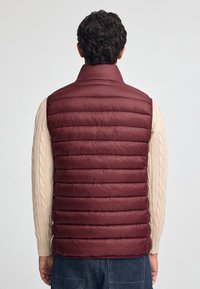 Chaleco tipo puff rojo burdeos con patrón de acolchado horizontal, cuello alto y material ligero, llevado sobre un suéter de punto en color crema.