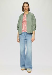 Hellgrüne Bomberjacke über einem rot-weiß gemusterten Hemd, kombiniert mit hellblauen, weiten Jeans und grünen Sneakers. Einfarbiger Hintergrund.