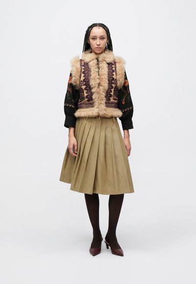 Αμάνικο γιλέκο από ψεύτικη γούνα σε μπεζ με περίπλοκη μοβ κέντημα, συνδυασμένο με φούστα χακί με φάσες. Σκούρα καλσόν και μπορντό ψηλοτάκουνα ολοκληρώνουν την εμφάνιση.