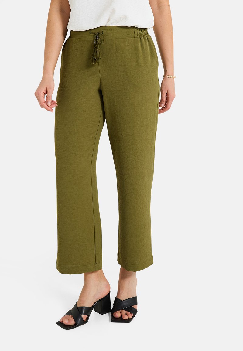 Pantalons larges verts olive en tissu texturé, avec taille élastique et cordon de serrage avec des glands, associés à des sandales à talons blocs noires.