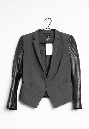 Grauer Blazer mit gesteppte schwarzen Ledermanschetten, Ein-Knopf-Verschluss und einem weißen rechteckigen Preisschild, das innen am Kragen hängt.