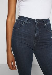 Mörkblå denim jeans med hög midja, dragkedjegylf, fem fickor och lätt blekning. Tyget har en slät yta och robust sömnad.