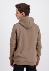 Ragazzino con capelli corti castani che indossa un semplice hoodie marrone e pantaloni neri, volto a sinistra su uno sfondo chiaro e uniforme.
