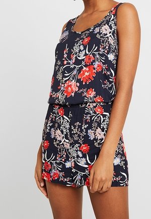 Granatowy kombinezon bez rękawów z czerwonym, białym i lawendowym kwiatowym wzorem, z elastyczną talią, noszony przez stojącą osobę z jedną zrelaksowaną ręką.