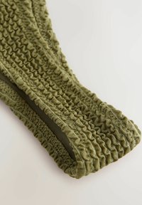 Olivgrön tyg med en texturerad, samlad design. Har elastiska kanter och ett slätt inre foder för komfort.