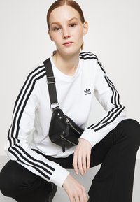 Baltas garās piedurknes krekls ar melnu Adidas logo un trīs melnām svītrām uz piedurknēm, apvienots ar melnām šaurām biksēm un mazu melnu rokassomu.