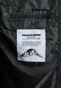 Črno najlonsko notranje oblogo zunanjega oblačila, ki vsebuje belo etiketo z besedilom "PEGADOR" in grafiko gore. Vključuje tudi žep z zadrgo.