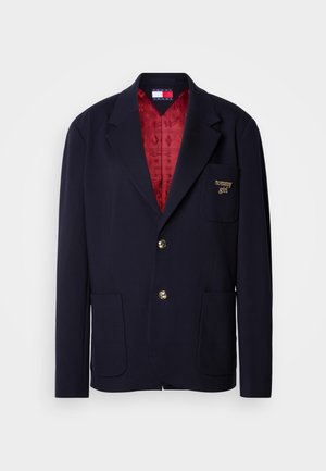 Mornarsko blazer iz mehkega blaga z rdečo vzorčasto podlogo. Ima dva sprednja žepa, zlato logo na prsih in zlato gumbanje.