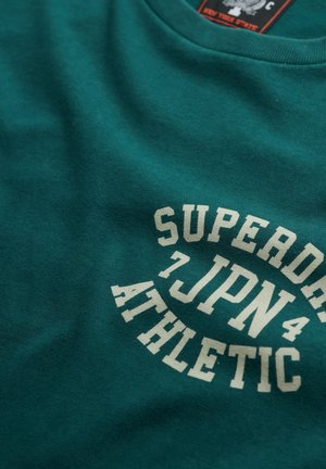 Teal bomulds T-shirt med rund halsudskæring og en stor hvid grafisk tekst, "SUPERDRY 7JPN ATHLETIC," i fed skrift.
