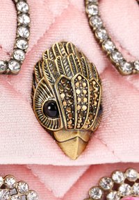 Brązowy broszka z misternym wzorem piór, czarnymi akcentami i błyszczącymi kryształkami, na tle teksturowanego różowego materiału.