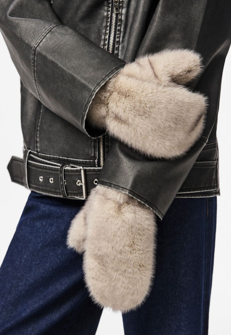 Luvas de pelo sintético em bege claro, com textura macia. Usadas com uma jaqueta de couro preta e jeans azuis, exibindo um design aconchegante.