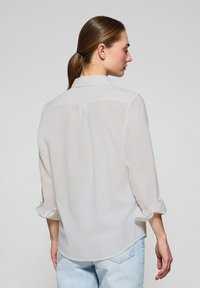 Biała, lekka koszula z guzikami z podwiniętymi rękawami, z detalem na plecach i zaokrąglonym hemem. Noszona z jasnoniebieskimi dżinsami.