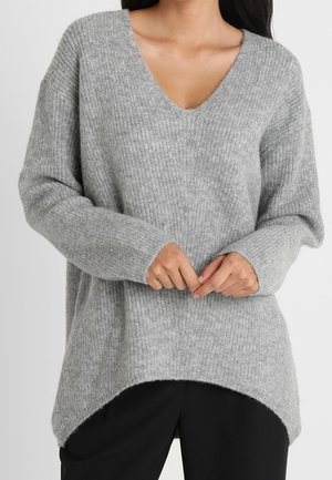 Femme portant un pull ample à manches longues en maille côtelée grise avec un col en V et un ourlet asymétrique, associé à un pantalon noir.