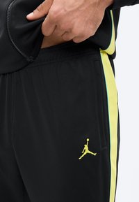 Zbliżenie dłoni podnoszącej czarną kurtkę, odsłaniając czarne sportowe spodnie z żółtymi bocznymi paskami i żółtym logo Jumpman w pobliżu kieszeni.